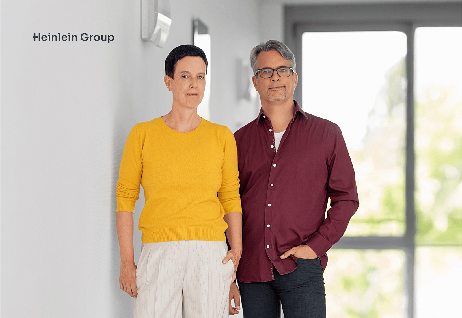 Jutta Horstmann und Peer Heinlein stehen nebeneinander in einem hellen Flur vor einem Fenster. Links ist das Logo ‚Heinlein Group‘ sichtbar.